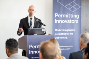 Jordan Eccles points toward the audience with a slide remote while speaking at the lectern.